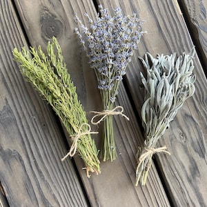 May include: Three bundles of dried herbs tied with twine on a wooden surface. The herbs include green thyme, purple lavender, and silvery sage. The bundles are arranged diagonally across the frame, showcasing their natural textures and colors.