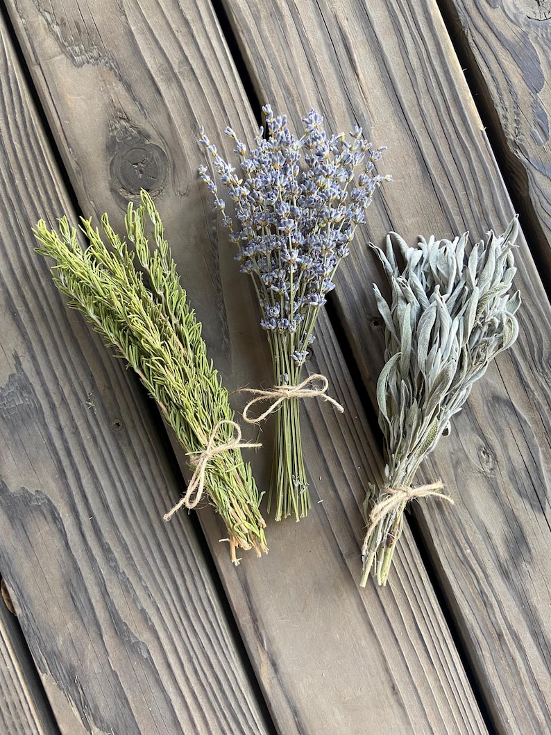 May include: Three bundles of dried herbs tied with twine on a wooden surface. The herbs include green thyme, purple lavender, and silvery sage. The bundles are arranged diagonally across the frame, showcasing their natural textures and colors.