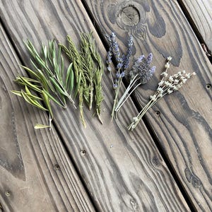 May include: An overhead shot of various dried herbs and flowers, including lavender, rosemary, and other green and beige foliage. The arrangement is set on a weathered wooden surface, with the natural colors of the plants contrasting against the wood.