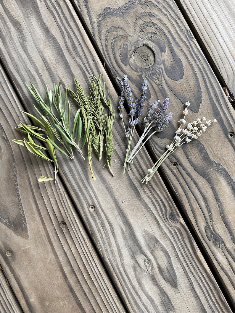 May include: An overhead shot of various dried herbs and flowers, including lavender, rosemary, and other green and beige foliage. The arrangement is set on a weathered wooden surface, with the natural colors of the plants contrasting against the wood.