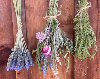 May include: Three bundles of dried herbs and flowers tied with twine. The left bundle features purple lavender. The center bundle contains a mix of lavender, a pink rose, and green foliage. The right bundle consists of green rosemary sprigs.