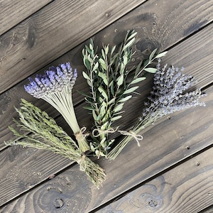 May include: An overhead shot of four bundles of dried herbs and flowers. The bundles include lavender, olive branches, and other green herbs, all tied with twine. The background is a dark wood surface.