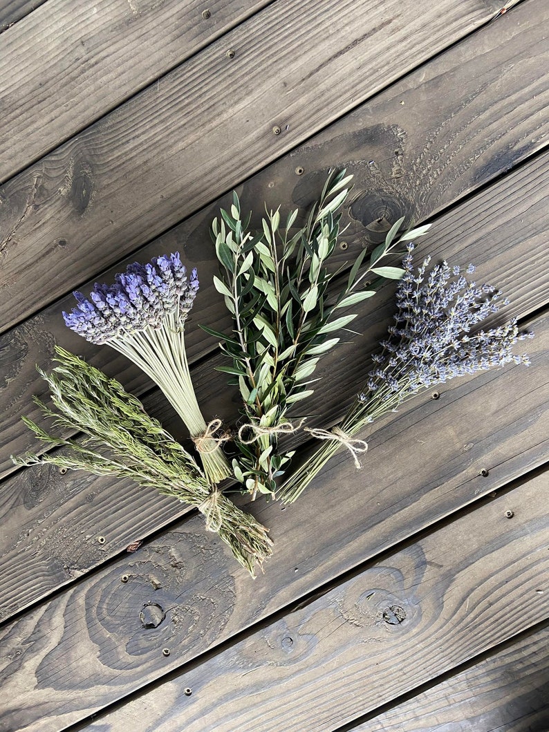 May include: An overhead shot of four bundles of dried herbs and flowers. The bundles include lavender, olive branches, and other green herbs, all tied with twine. The background is a dark wood surface.
