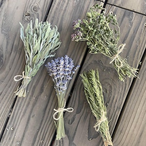 May include: Four bundles of dried herbs tied with twine, including sage, lavender, oregano, and rosemary. The herbs are arranged on a weathered, gray wooden surface, showcasing their natural colors and textures. The bundles are tied with natural twine.