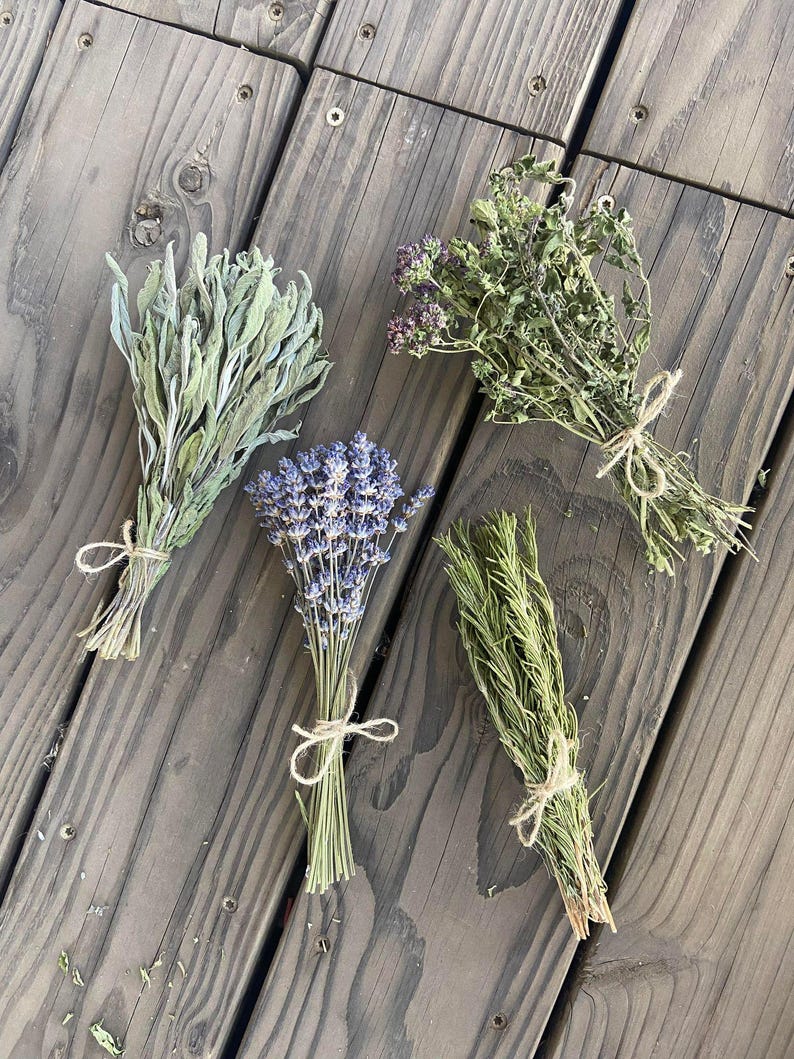 May include: Four bundles of dried herbs tied with twine, including sage, lavender, oregano, and rosemary. The herbs are arranged on a weathered, gray wooden surface, showcasing their natural colors and textures. The bundles are tied with natural twine.