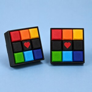 May include: Two square black frames each containing a three-by-three grid of colorful plastic keycaps. The top rows are red, orange, and yellow. The middle rows are black with a red heart icon in the center. The bottom rows are green, blue, and dark blue.
