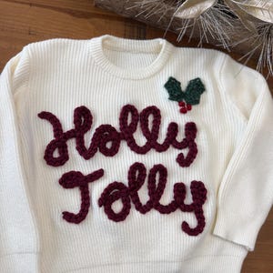 May include: White knitted sweater with a red embroidered "Holly Jolly" design and a green holly sprig with red berries.