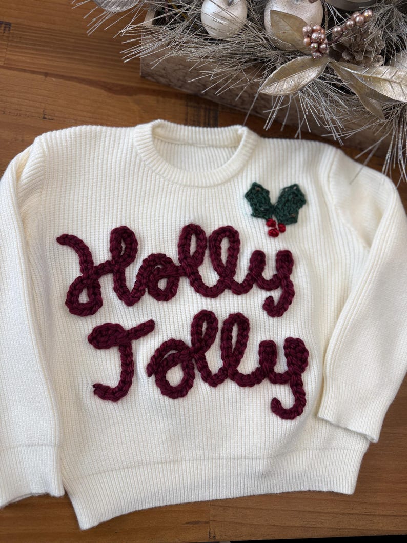 May include: White knitted sweater with a red embroidered "Holly Jolly" design and a green holly sprig with red berries.