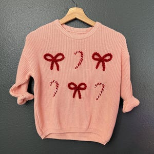 May include: A pink knitted sweater with red bows and candy cane designs. The sweater has a crew neck and long sleeves.