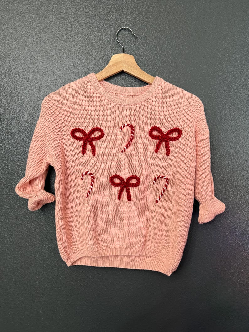 May include: A pink knitted sweater with red bows and candy cane designs. The sweater has a crew neck and long sleeves.