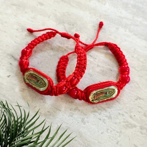 May include: Two red woven bracelets with a religious image in the center. The bracelets have red beads and are displayed on a light gray surface with green foliage in the corner.