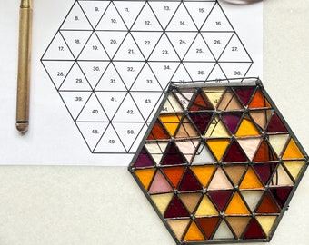Könnte beinhalten: Ein sechseckiges Glasmosaik mit verschiedenen Farben wie Rot, Orange, Gelb und Braun. Das Sechseck besteht aus vielen kleinen Dreiecken. Im Hintergrund ist ein Blatt Papier mit einem Raster aus nummerierten Dreiecken zu sehen. Ein Rolle Kupferklebeband ist ebenfalls im Hintergrund zu sehen.