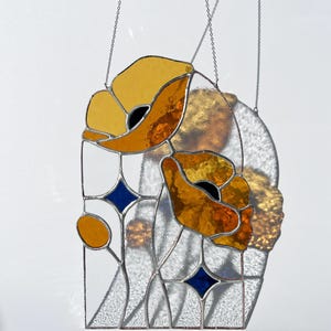 May include: Stained glass panel featuring two stylized orange and yellow poppy flowers with black centers. The design includes blue diamond accents and is framed with silver solder. The panel hangs from a silver chain.