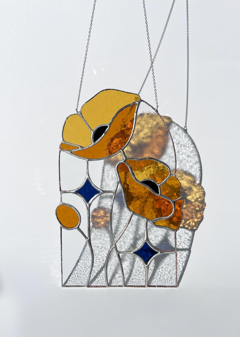 May include: Stained glass panel featuring two stylized orange and yellow poppy flowers with black centers. The design includes blue diamond accents and is framed with silver solder. The panel hangs from a silver chain.