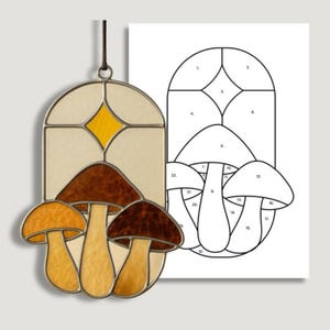 May include: A stained glass mushroom suncatcher with a brown, yellow, and cream color scheme. The suncatcher is hanging next to a black and white pattern for the stained glass design.