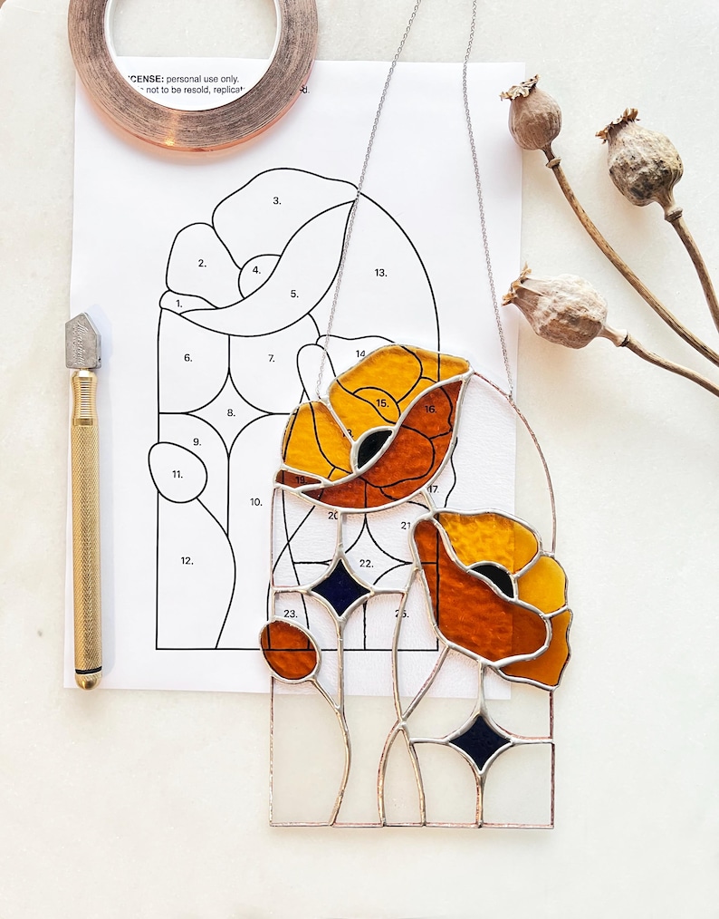 May include: A stained glass poppy art piece with orange and yellow petals, black centers, and clear glass stems. A pattern, glass cutter, copper tape, and dried seed pods are also present.