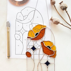 May include: A stained glass poppy art piece with orange and yellow petals, black centers, and clear glass stems. A pattern, copper tape, and a glass cutter are nearby. The text on the pattern reads "LICENSE: personal use only. not to be resold, replica."