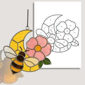 May include: Stained glass suncatcher featuring a bee, a crescent moon, and a pink flower. The bee has yellow, brown, and white sections. The moon is yellow. A paper with a flower pattern is in the background.
