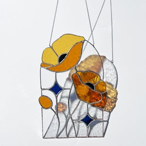 May include: Stained glass panel featuring two stylized orange and yellow poppies with black centers. The design includes blue diamond shapes and clear glass, all framed with a metal border and suspended by chains. A decorative floral piece.