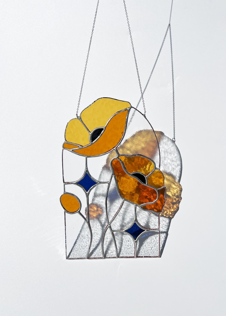 May include: Stained glass panel featuring two stylized orange and yellow poppies with black centers. The design includes blue diamond shapes and clear glass, all framed with a metal border and suspended by chains. A decorative floral piece.