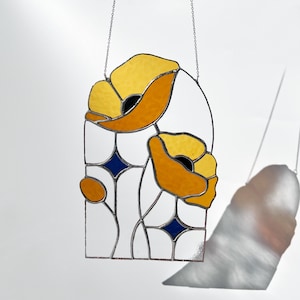 May include: Stained glass window panel featuring stylized yellow and orange poppies with black centers. The design includes white and blue geometric shapes, all framed by a black border. The panel is suspended by a silver chain.