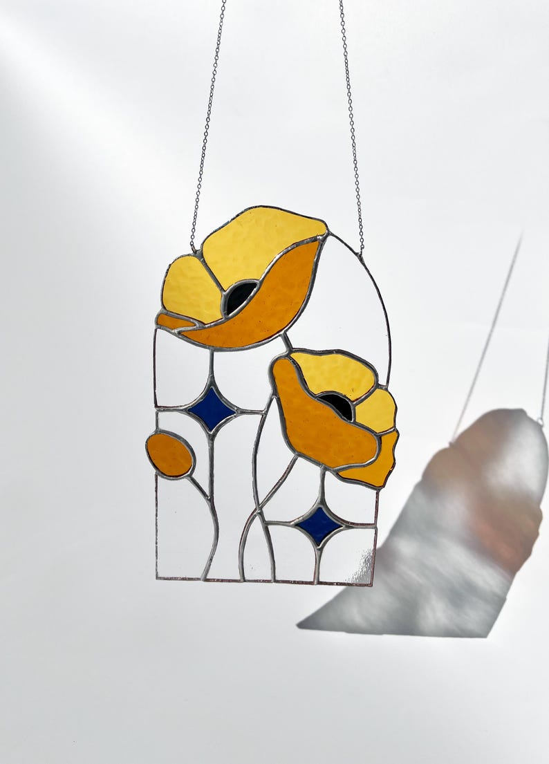 May include: Stained glass window panel featuring stylized yellow and orange poppies with black centers. The design includes white and blue geometric shapes, all framed by a black border. The panel is suspended by a silver chain.