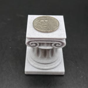May include: A small, light gray miniature Ionic column with a coin on top. The column has detailed carvings and a square base. The coin is silver-colored and features a portrait.