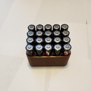 May include: A brown plastic battery storage box with the label "AA" on the front. The box is filled with 24 AA batteries.