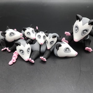May include: A group of six gray and white opossum figurines with pink noses and black eyes. The opossums are in various poses, some are lying down, some are standing up, and some are sitting. The figurines are made of a smooth, plastic-like material.