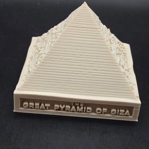 May include: A beige model of the Great Pyramid of Giza, with the text "THE GREAT PYRAMID OF GIZA" at the base. The pyramid has a textured surface and is set against a dark background. The model is a detailed replica.