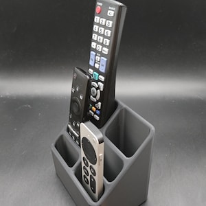 May include: A gray plastic remote control organizer holding three different remote controls. The remotes are black and silver, with various buttons and labels. The organizer has multiple compartments.