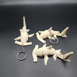 May include: Three white plastic hammerhead shark keychains. Each keychain is articulated and can be posed in different positions. The keychains are attached to silver key rings.
