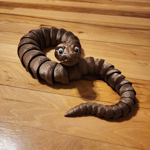 May include: A brown segmented snake toy with large eyes and a toothy grin. The snake is curled up on a wooden floor.