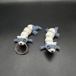 May include: Two gray and white ferret figurines with black eyes. One ferret has a gold ring attached to its body.