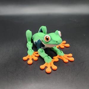 May include: A green and orange 3D printed frog figurine with articulated limbs. The frog has large, black eyes with orange pupils.