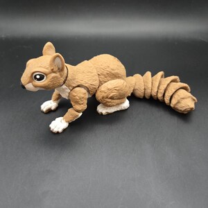 May include: A brown articulated squirrel figurine with a segmented tail. The toy squirrel has white paws and a white chest. The squirrel has a black collar and large, expressive eyes. The figurine is posed on a dark surface.