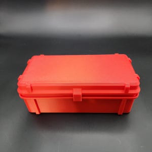Puede incluir: Una caja de almacenamiento de pl&aacute;stico roja con una tapa con bisagras. La caja es rectangular y tiene una superficie lisa.