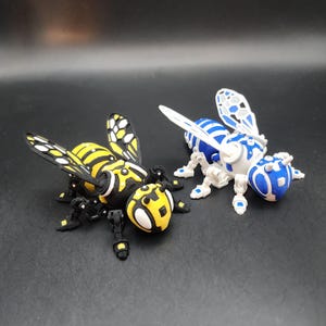 May include: Two 3D printed robotic bee toys, one black and yellow, the other white and blue. Both have articulated legs and wings.