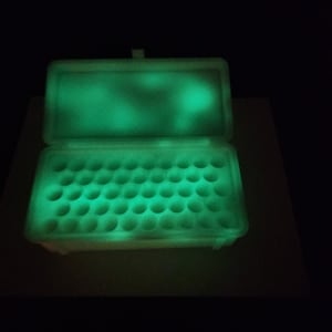 Puede incluir: Un contenedor de pl&aacute;stico blanco con tapa, que brilla en verde en la oscuridad. El contenedor tiene m&uacute;ltiples compartimentos circulares para guardar objetos peque&ntilde;os.
