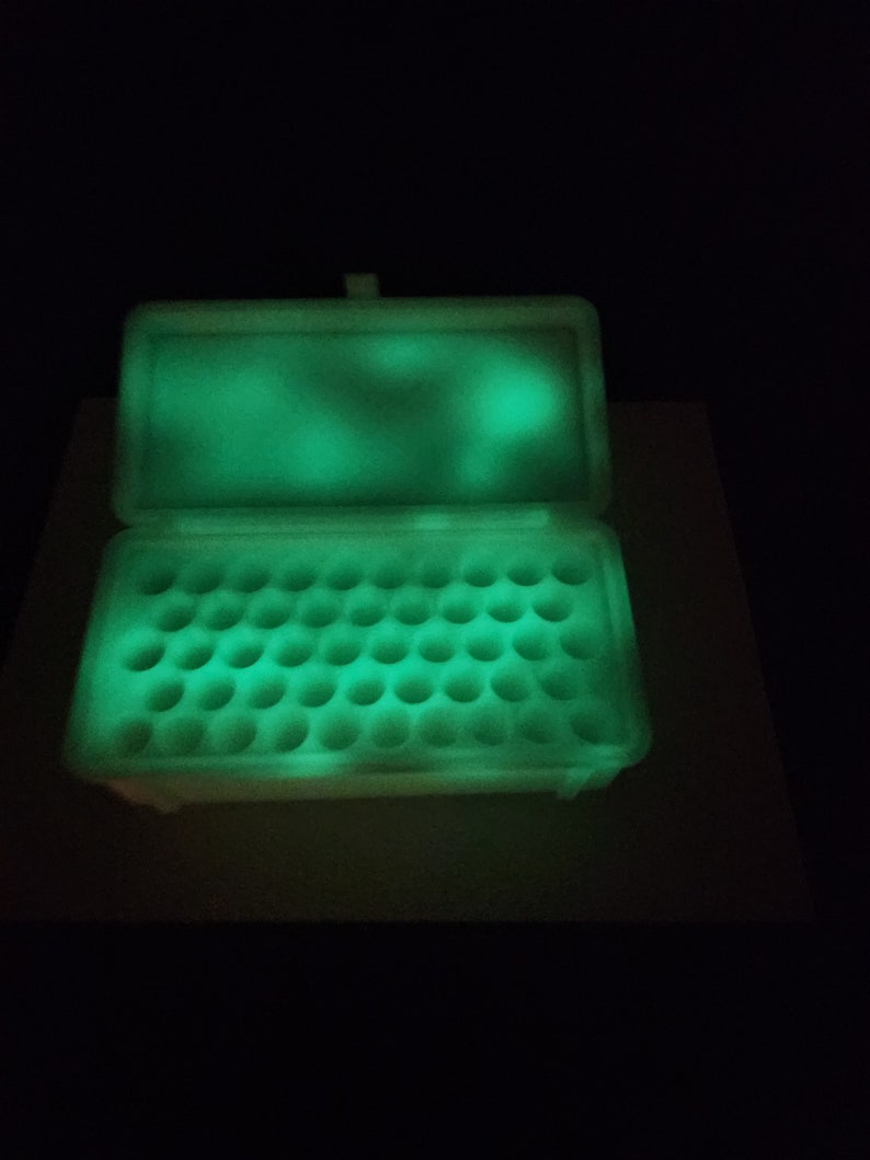 Puede incluir: Un contenedor de pl&aacute;stico blanco con tapa, que brilla en verde en la oscuridad. El contenedor tiene m&uacute;ltiples compartimentos circulares para guardar objetos peque&ntilde;os.