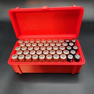 Puede incluir: Una caja de almacenamiento de pl&aacute;stico roja llena de bater&iacute;as de diferentes tama&ntilde;os. La caja est&aacute; abierta y las bater&iacute;as son visibles. Hay bater&iacute;as AA y AAA en la caja.