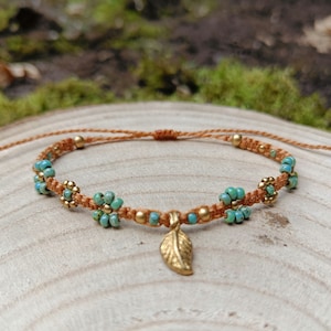 Puede incluir: Pulsera de macramé hecha a mano con cuentas de flores turquesas y detalles dorados. La pulsera tiene un dije de hoja dorada y se exhibe sobre una superficie de madera. Accesorio de estilo bohemio, ajustable.