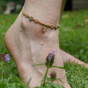 以下が含まれることがあります： 女性の足首のクローズアップで、小さな金のチャームが付いた繊細なアンクレットをしています。アンクレットは茶色の紐で作られており、小さなターコイズビーズが付いています。足首は緑の草と紫の花に囲まれています。