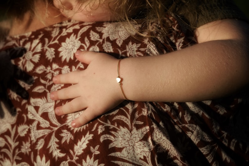 Może przedstawiać: Delikatna bransoletka z zawieszką w kształcie złotego serca na nadgarstku dziecka. Bransoletka posiada małą zawieszkę w kształcie serca na cienkim, brązowym sznurku. Ramię dziecka widoczne jest na tle tkaniny w brązowo-biały wz&oacute;r kwiatowy.
