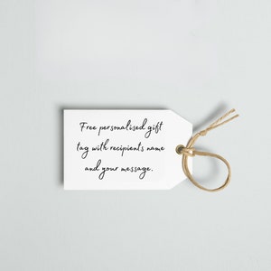 Może przedstawiać: Biała etykieta prezentowa z napisem "Free personalised gift tag with recipient's name and your message" czcionką w kolorze czarnym. Brązowy sznurek jest przełożony przez złoty otw&oacute;r u g&oacute;ry etykiety.