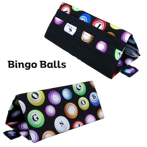 Pu&ograve; includere: Due porta-biglie da bingo in tessuto nero con un motivo colorato di biglie da bingo. I porta-biglie sono di forma triangolare e presentano la scritta "Bingo Balls". Le biglie da bingo sono di vari colori, tra cui rosso, verde e viola.