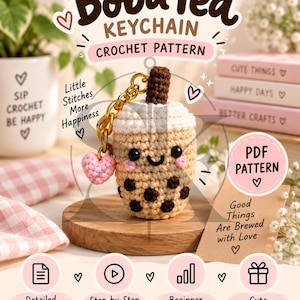 May include: A crochet boba tea keychain pattern featuring a tan cup with dark brown tapioca pearls, a white foam top, and a brown straw. The design includes a small pink heart charm, a gold metal keyring, and text reading Boba Tea Keychain Crochet Pattern.