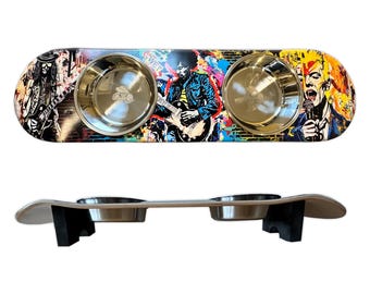 May include: A dog bowl set made from a repurposed skateboard. The skateboard has a colorful graphic design featuring musicians. Two stainless steel bowls are set into the skateboard. The skateboard is supported by two black wooden legs.