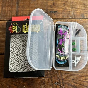 Puede incluir: Una caja de plástico transparente con un fingerboard en su interior. El fingerboard tiene un gráfico de calavera colorido. La caja tiene compartimentos para herramientas y otros accesorios. La caja está etiquetada como "Fingerboards".