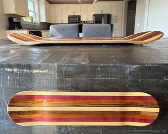 May include: Two wooden skateboards with a striped design. The boards are made of various wood tones, including light brown, red, and purple. The boards are displayed on a dark wooden table.